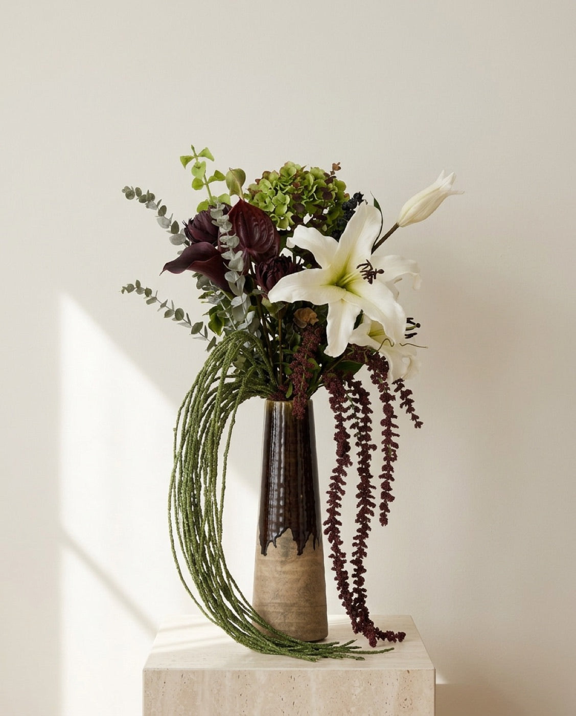 Composition Blooma : lys blanc, anthurium bordeaux, hortensia vert et amarante retombante dans un vase haut émaillé brun, posé sur un socle en travertin dans une lumière de fin de journée.
